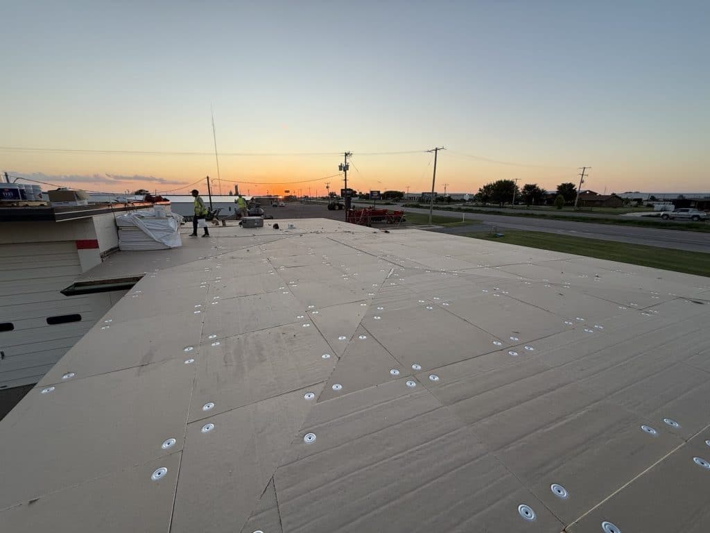 During-restoration photo of a commercial flat roof with new tan/beige insulation cover board mechanically fastened over the existing substrate. Numerous rows of large white screw plates and fasteners are visible across the freshly installed rigid ISO cover board. A worker in a high-visibility vest stands near stacked materials at sunset, with a highway and industrial buildings in the background.